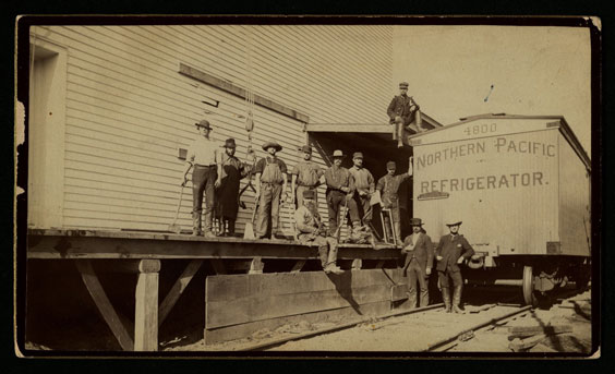 Men stand and sit on a platform next to a train car that reads Northern Pacific Refrigerator. Two men stand in front of the train car.